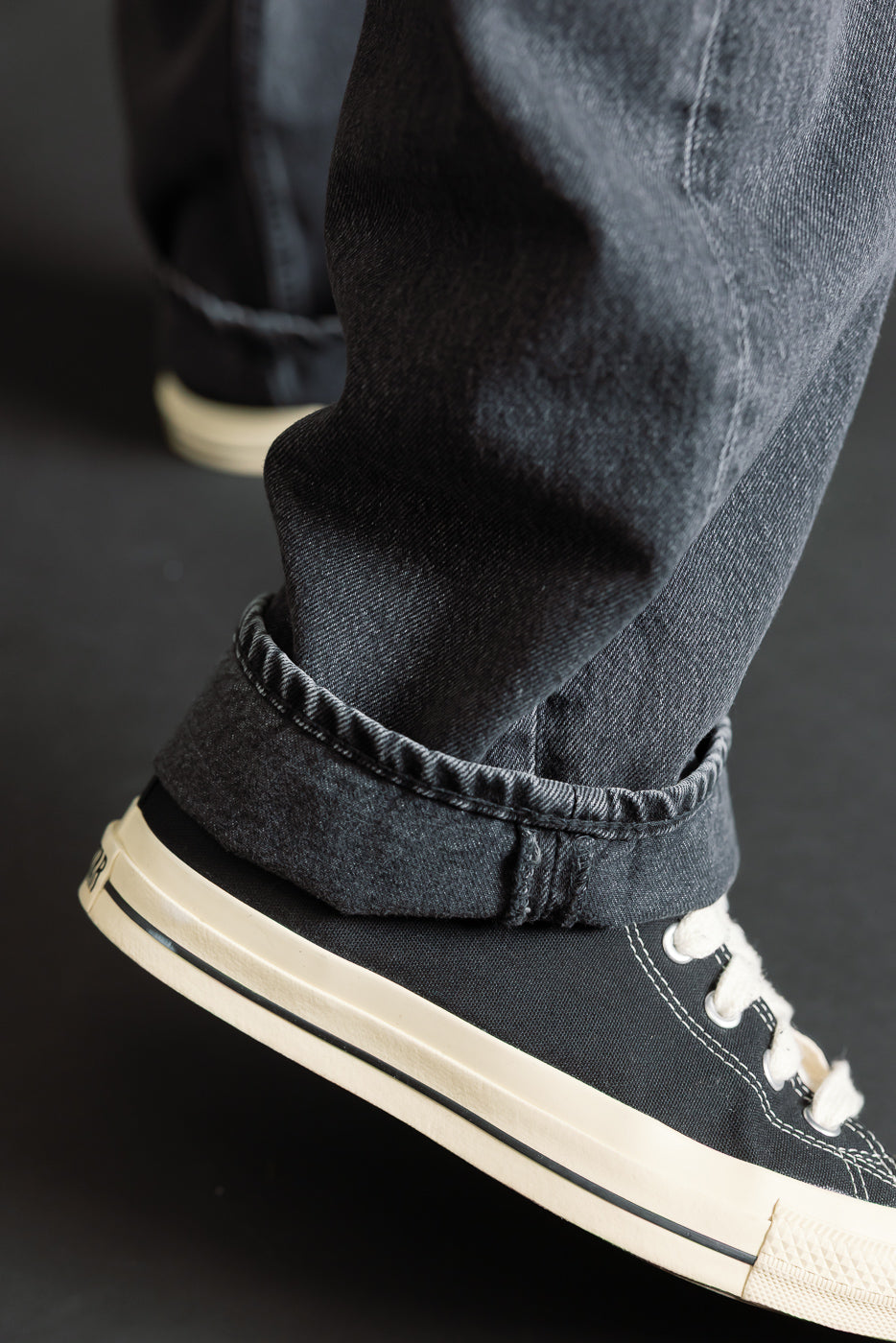Close-up of a lower leg in 01-1010W-D61S - 101 Dad’s Fit Denim - 90s Black Stone, showcasing relaxed black stone-washed denim with a cuffed hem, paired with black canvas sneakers.