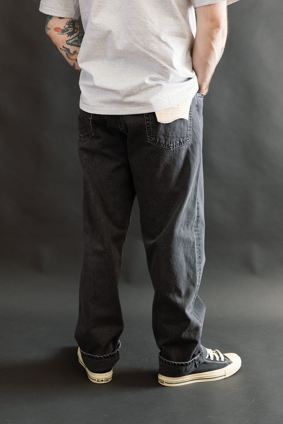 A man wearing 01-1010W-D61S - 101 Dad’s Fit Denim - 90s Black Stone jeans, showcasing a relaxed, vintage-inspired fit with black denim and a distinctive tattoo on his left forearm, styled with sneakers.