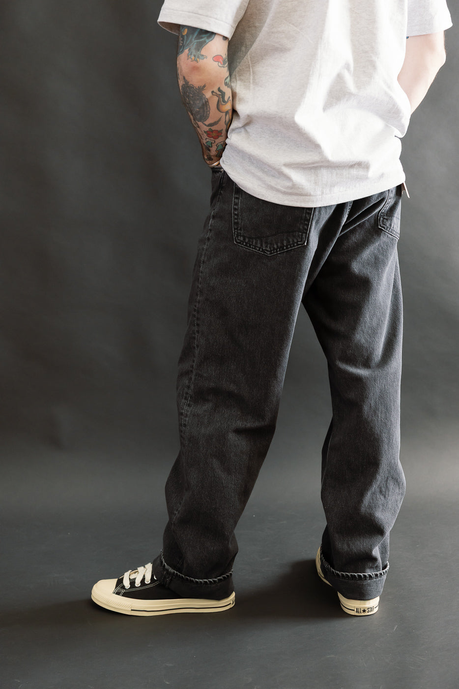 Person wearing 01-1010W-D61S - 101 Dad’s Fit Denim - 90s Black Stone, showcasing loose-fit black stone-washed jeans with rolled cuffs, paired with black canvas sneakers and visible tattooed forearm.