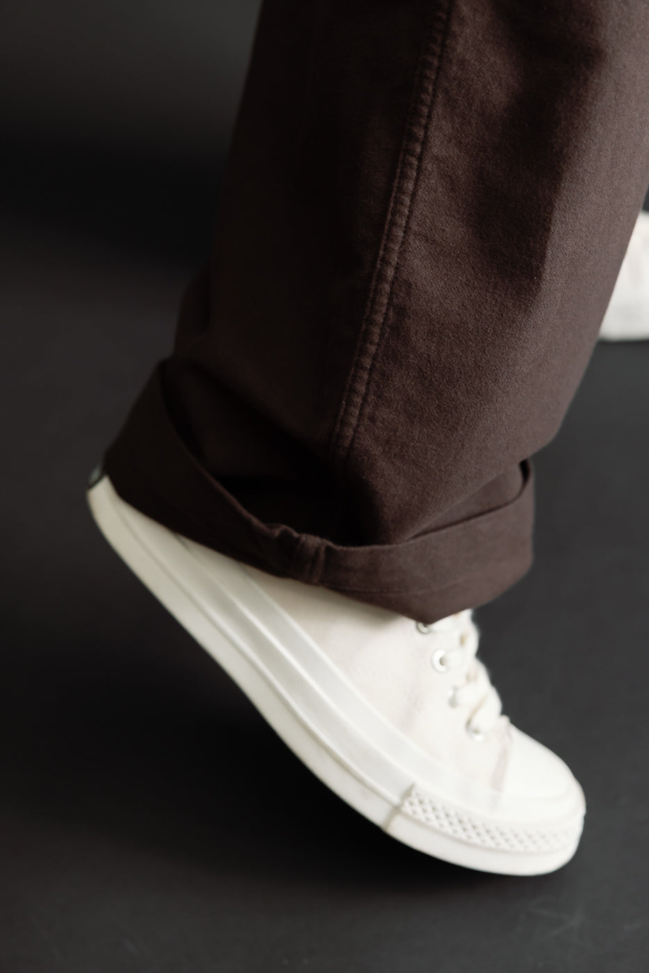 Close-up of the cuffed hem of brown trousers and white canvas sneakers on a male model.