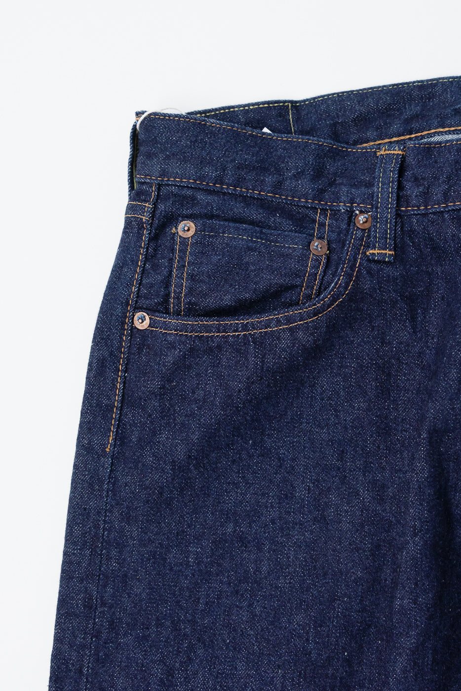 Close-up of front left waist and pocket of orSlow 1950s selvedge denim jeans, showing raw indigo fabric, orange contrast stitching, rivet detailing, and belt loop, flat lay.