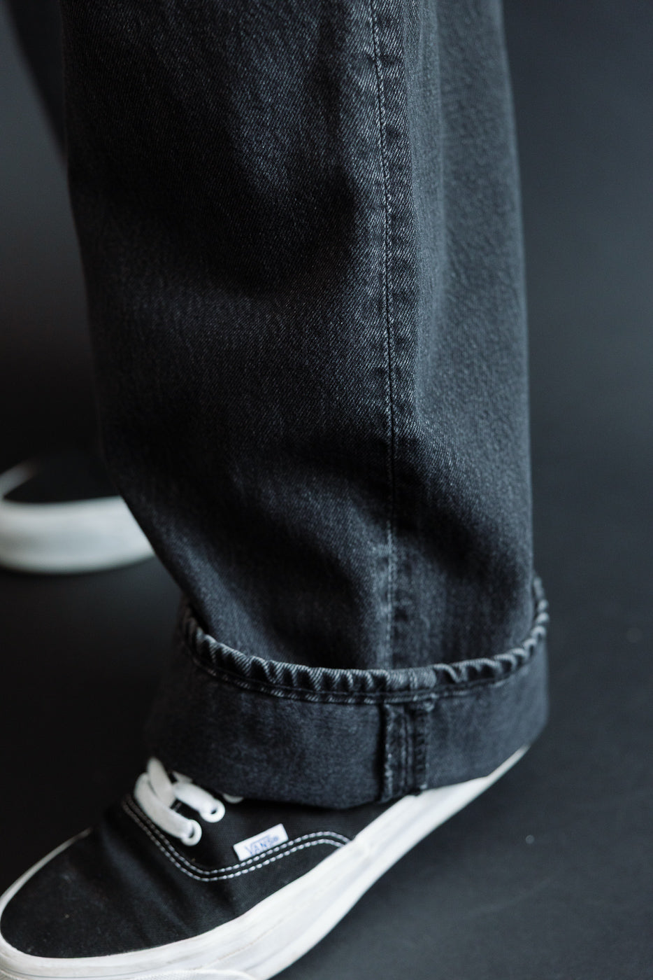 Close-up of male model's lower leg in dark selvedge jeans with cuffed hem, paired with black Vans sneakers