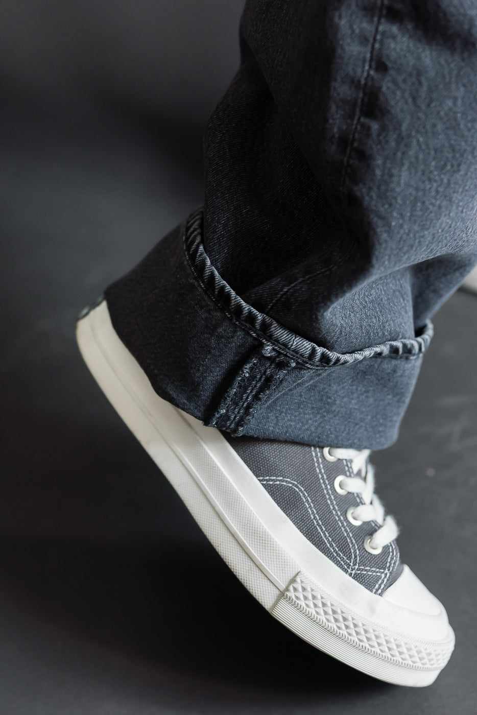 Close-up of a male model's foot wearing grey canvas sneakers and dark denim jeans with a cuffed hem.