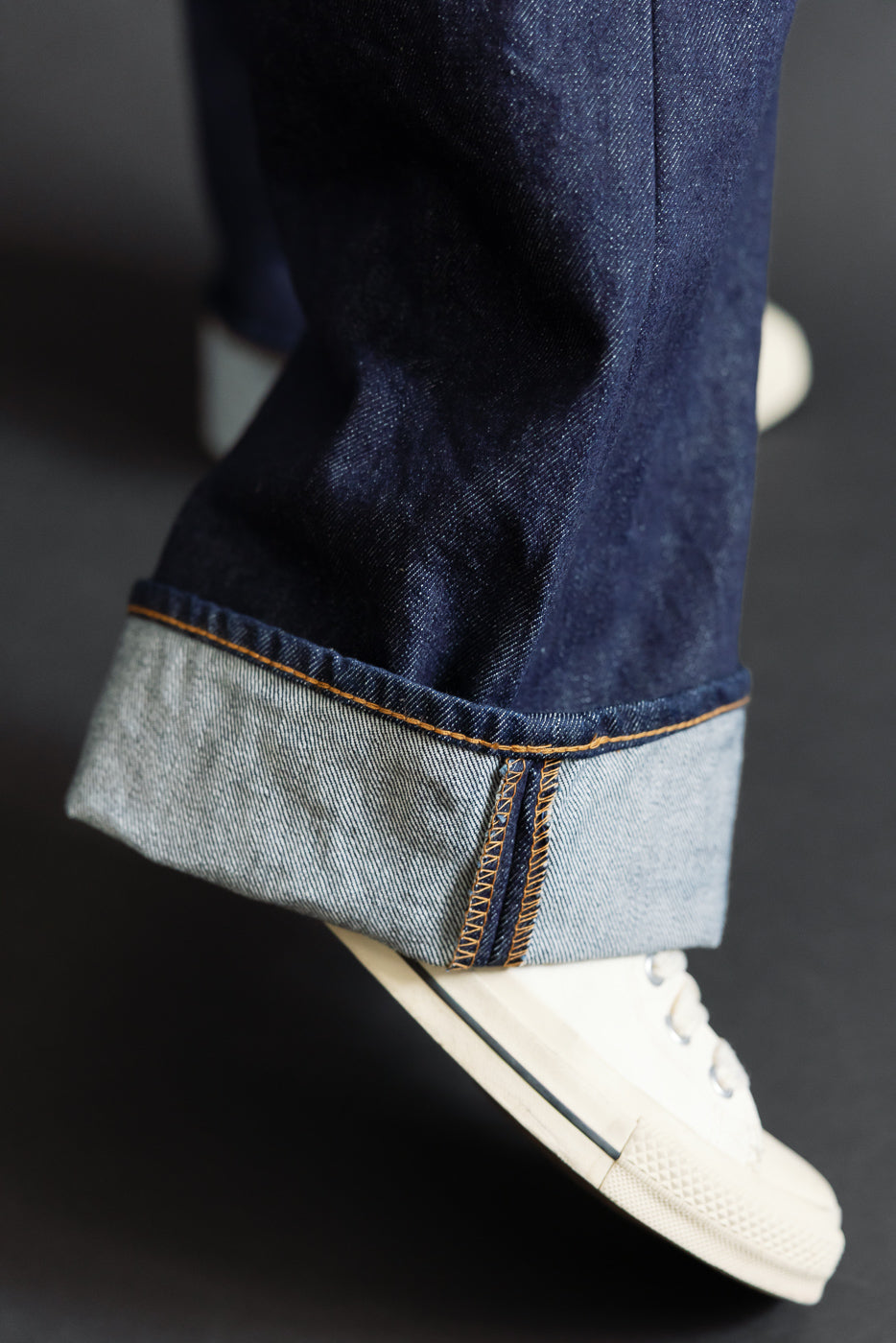 Close-up of the cuffed hem and selvedge detail on dark indigo denim jeans worn by a male model.