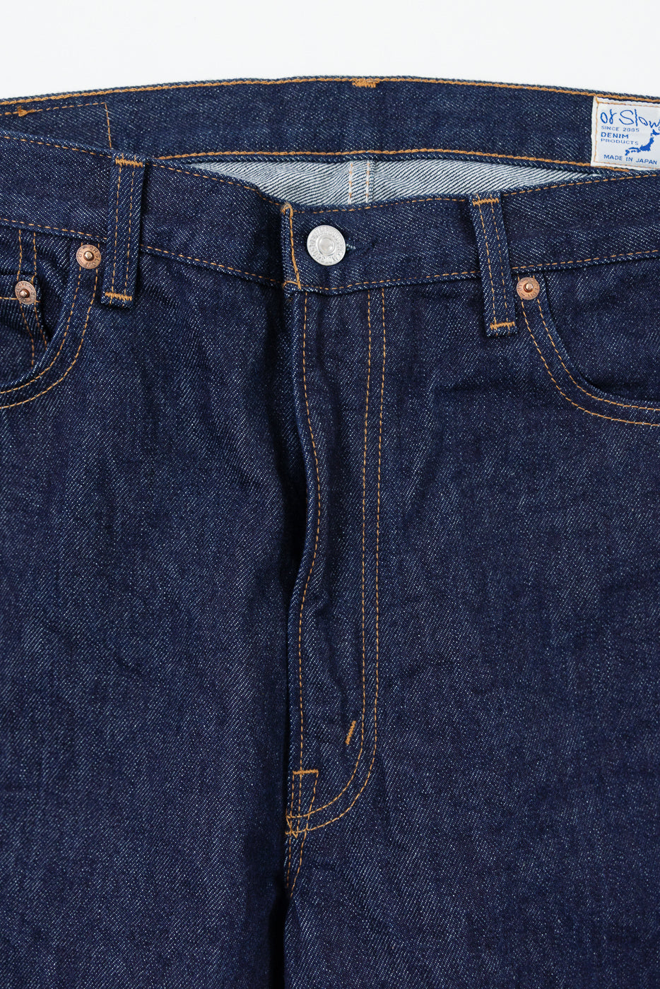 Close-up front view of dark indigo selvedge denim jeans showing the waistband, button, and stitching.