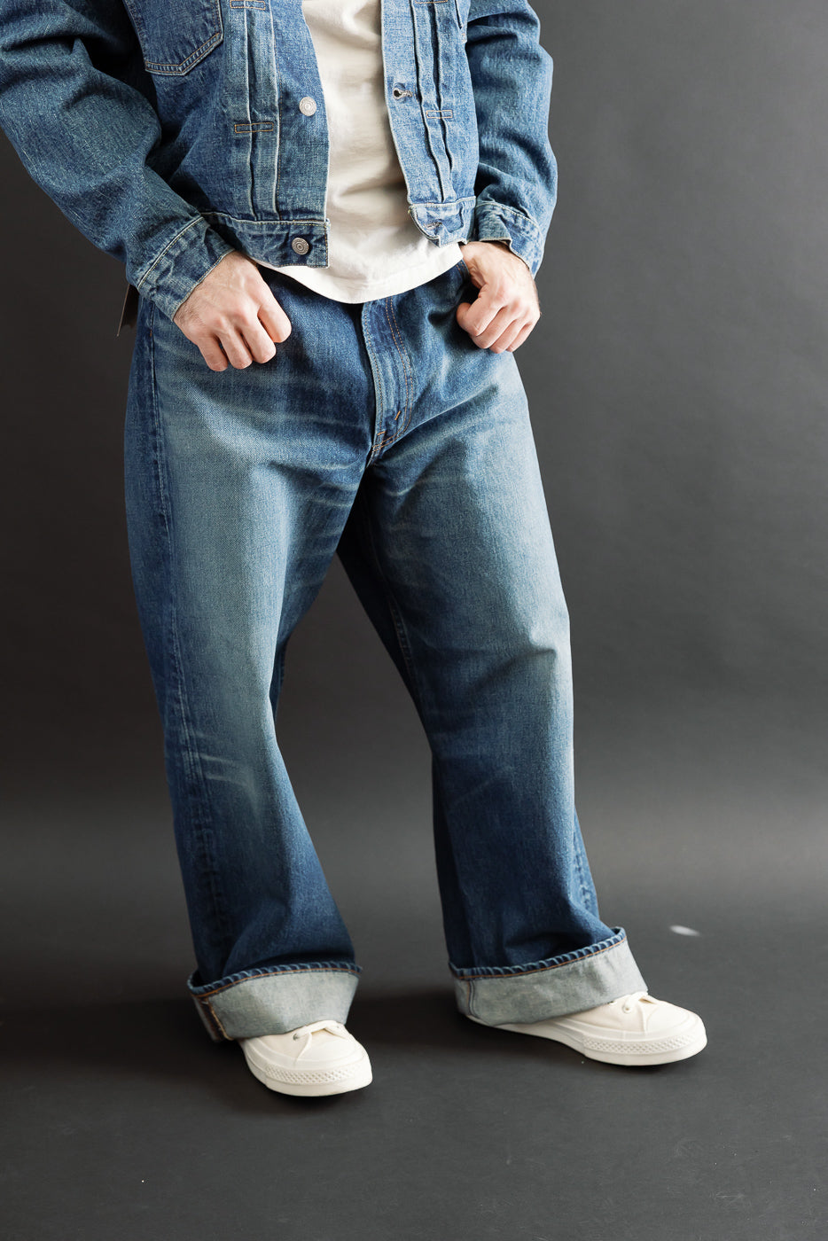 Full body front view of a male model with an athletic build wearing baggy blue denim jeans and a denim jacket.