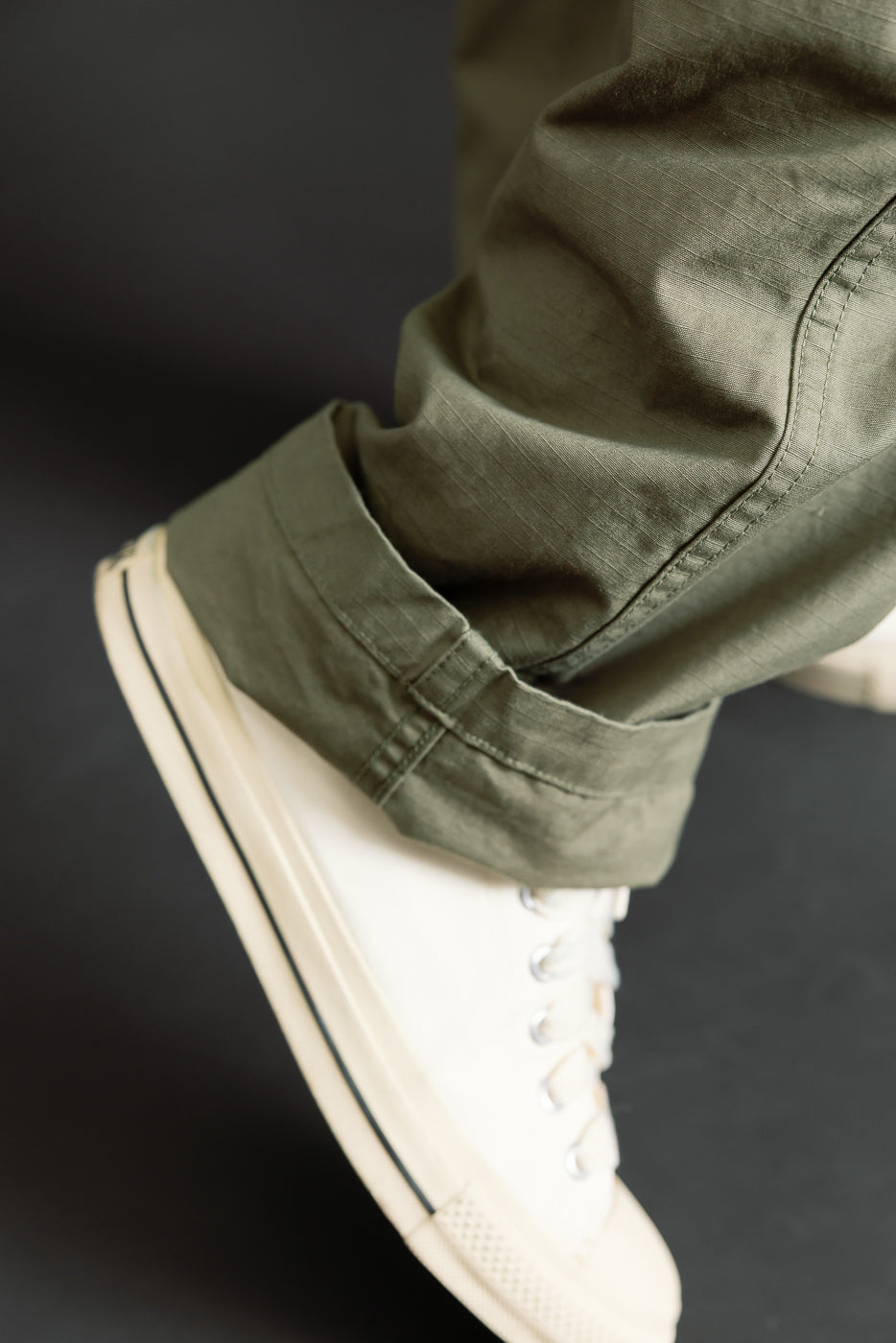 Close-up of men's fatigue pants cuff in ripstop army green, front view, on athletic male model, showing fabric texture next to white sneaker.