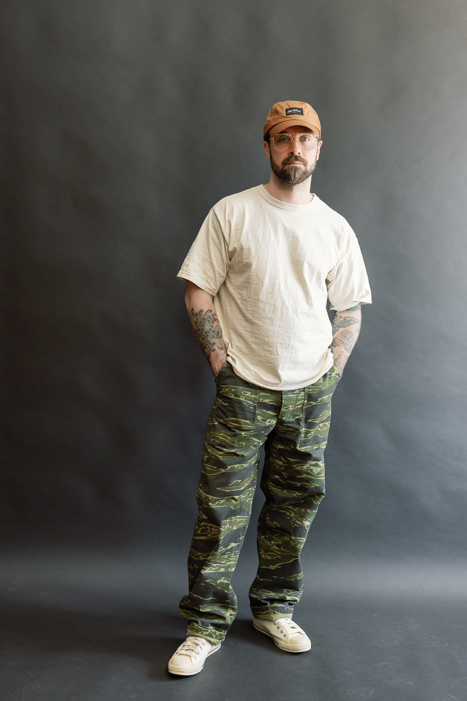 Man wearing 01-5002-TGC Fatigue Pants Ripstop Standard Fit in Tiger Camo with a light-beige T-shirt, off-white sneakers, and a tan cap, showcasing a relaxed, heritage-inspired ensemble.