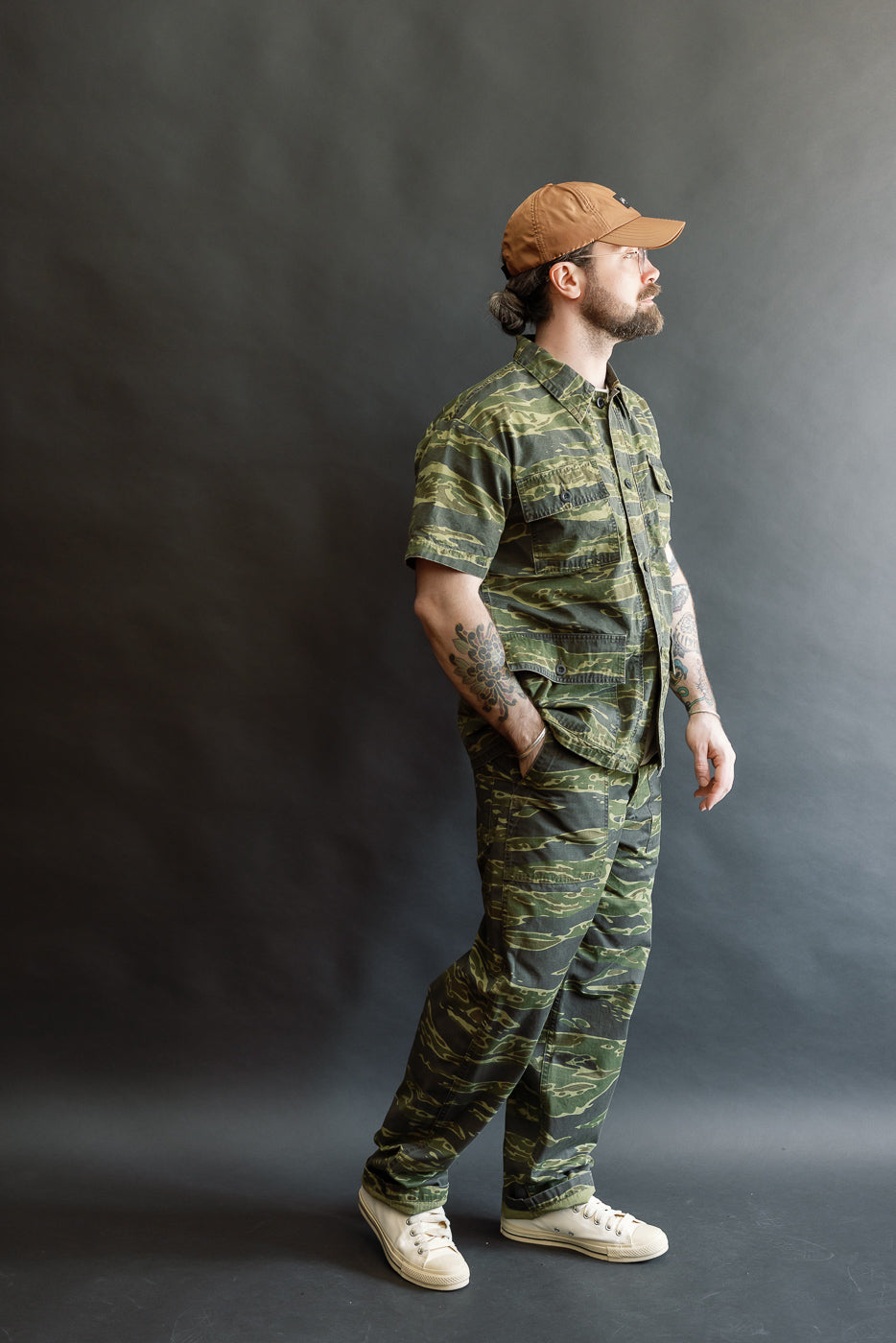A bearded man models orSlow 01-5002-TGC Fatigue Pants Ripstop Standard Fit - Tiger Camo, showcasing their straight fit, tiger camo pattern, and side-tab waist adjustments, paired with matching shirt, sneakers, and cap.