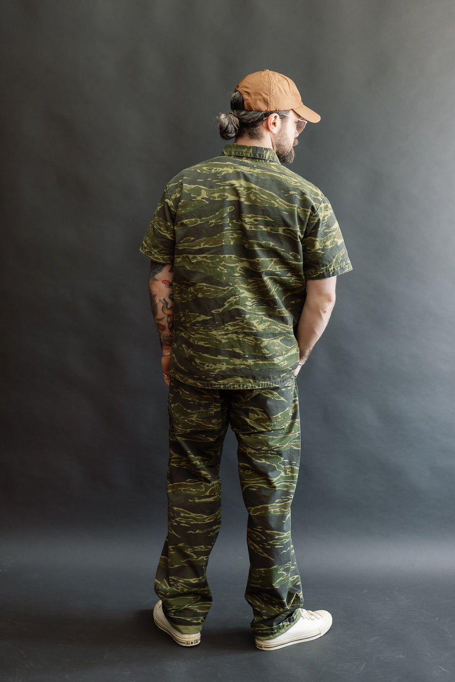 A man models the orSlow 01-5002-TGC Fatigue Pants Ripstop Standard Fit in Tiger Camo, with matching shirt and off-white sneakers. His back is turned, displaying tattoos and a low bun.