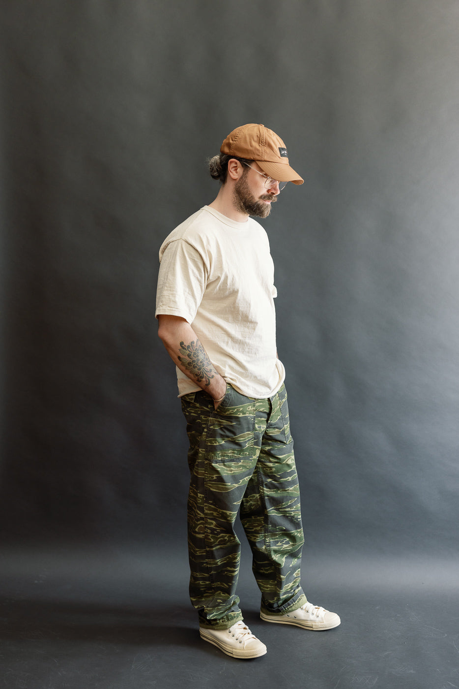 A bearded man models the orSlow 01-5002-TGC Fatigue Pants in tiger camo ripstop, showcasing their standard fit and durability, styled with a T-shirt and sneakers in a studio setting.