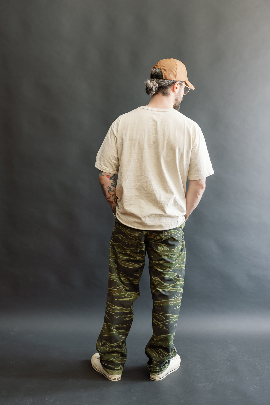 Man in 01-5002-TGC Fatigue Pants Ripstop Standard Fit - Tiger Camo, showcasing straight fit camouflage pants with side-tab waist adjustments, paired with a beige tee and canvas sneakers, standing relaxed.