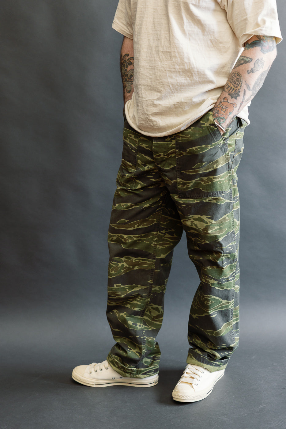 The 01-5002-TGC Fatigue Pants Ripstop Standard Fit - Tiger Camo feature large patch pockets and a straight fit, showcased with rolled-up sleeves and canvas sneakers against a gray backdrop.