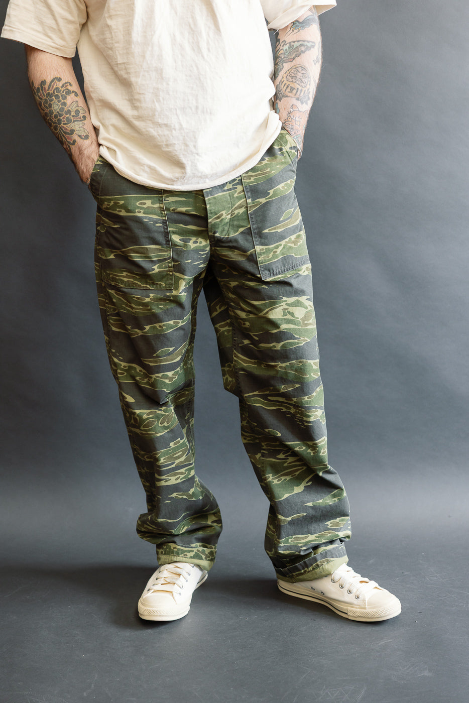 Model wearing orSlow 01-5002-TGC Fatigue Pants Ripstop Standard Fit - Tiger Camo, showcasing loose-fitting camouflage cargo pants with side-tab waist adjustments and period-correct buttons, paired with casual sneakers.