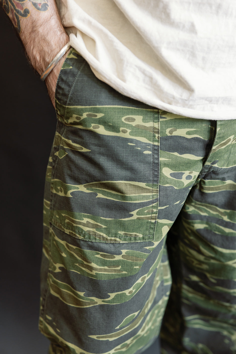 01-5002-TGC - Fatigue Pants Ripstop Standard Fit - Tiger Camo, featuring green tiger-stripe camouflage, shown worn with hand in pocket, showcasing ripstop texture and silver bangle accessory.