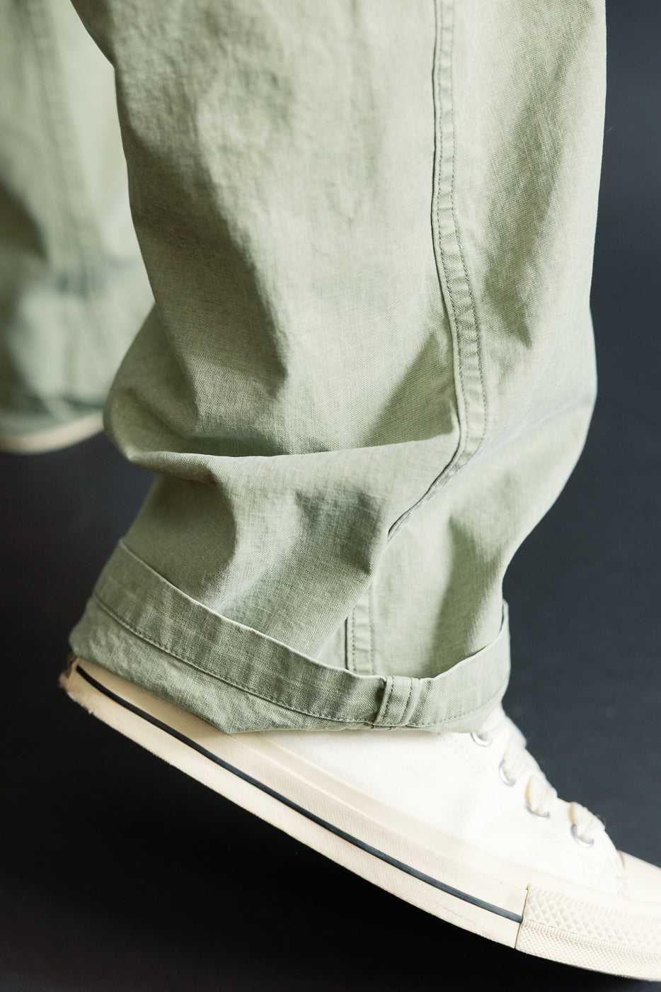 Close-up of male model's lower leg in olive green casual pants with cuffed hem, paired with white sneakers