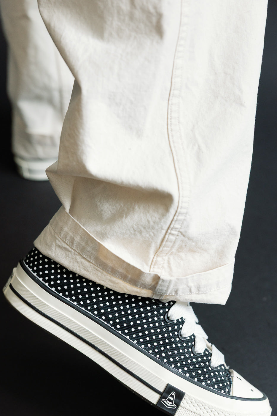 Close-up of male model’s lower leg in off-white pants with cuffed hem, paired with black polka dot sneakers, side view