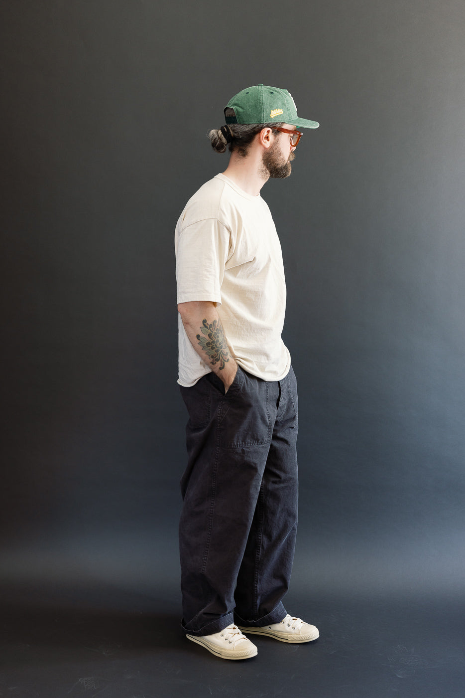 A man models the orSlow 01-5103-M61K Duck Canvas Summer Fatigue Pants in black, showcasing their high-rise, wide-leg fit and functional deep pockets, styled with casual attire.