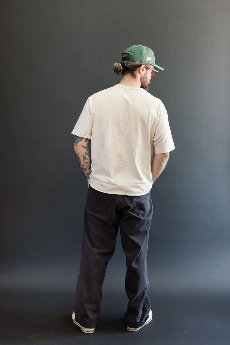 Man wearing orSlow 01-5103-M61K Duck Canvas Summer Fatigue Pants - Black, styled with a light-beige T-shirt, white sneakers, and a green cap, highlighting the pants' high rise and roomy fit.