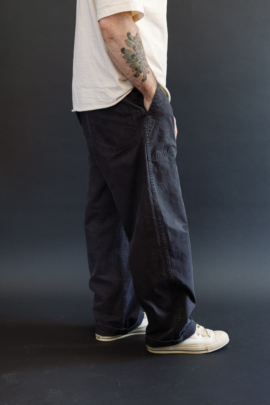 Person wearing 01-5103-M61K - Duck Canvas Summer Fatigue Pants - Black, showcasing a high-rise, wide-leg fit with hand in pocket, paired with off-white canvas sneakers and a visible floral forearm tattoo.