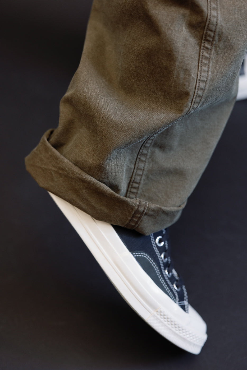 Close-up side view of olive selvedge pants with cuffed hem, worn by male model, paired with black canvas sneakers