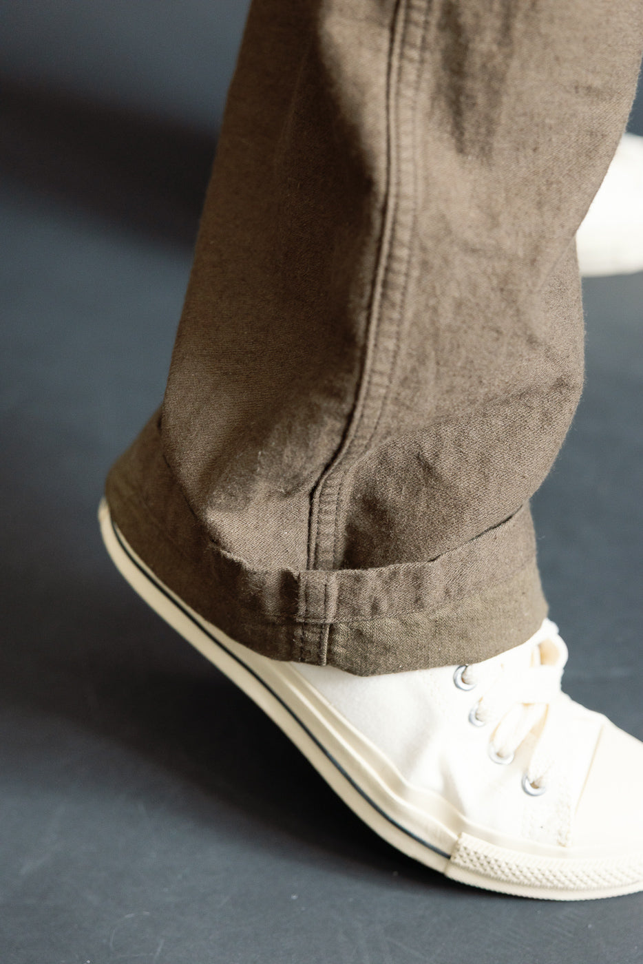 Close-up of the front lower hem of orSlow coffee brown fatigue pants, cotton-linen, on male model, showing fabric texture and straight hem, styled with white sneakers.