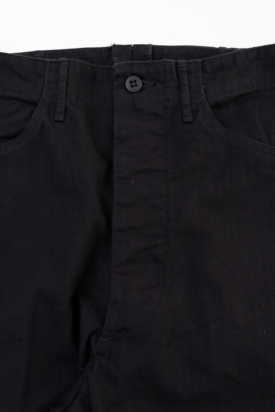 Close-up front view of black cotton trousers showing the waistband, button, and fly detail.