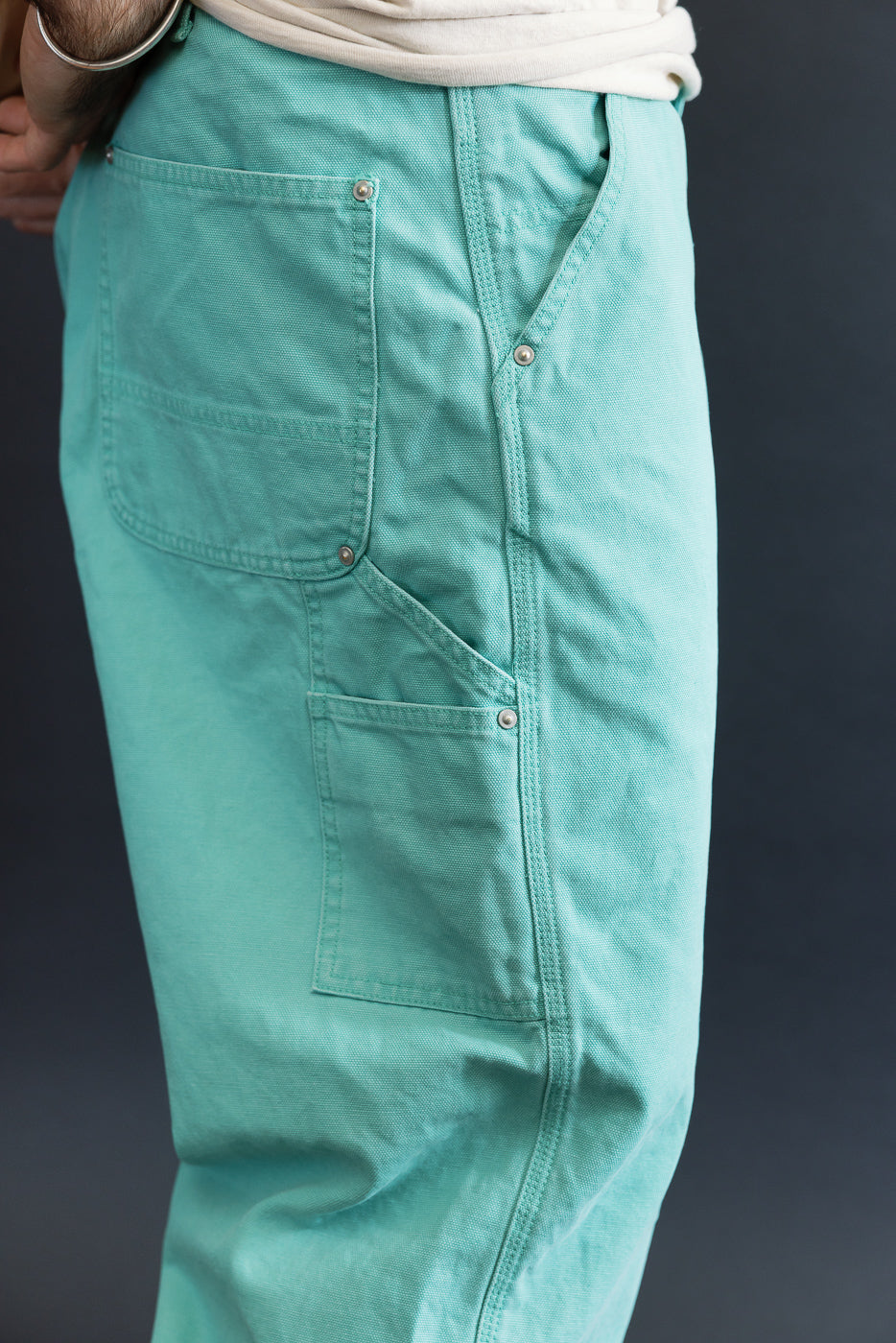 Close-up of 01-5328-39SK - Dad's Fit Painter's Pants Canvas - Turquoise, showcasing large patch pockets with brass UFO rivet details, styled with a tucked-in light-colored shirt against a dark backdrop.