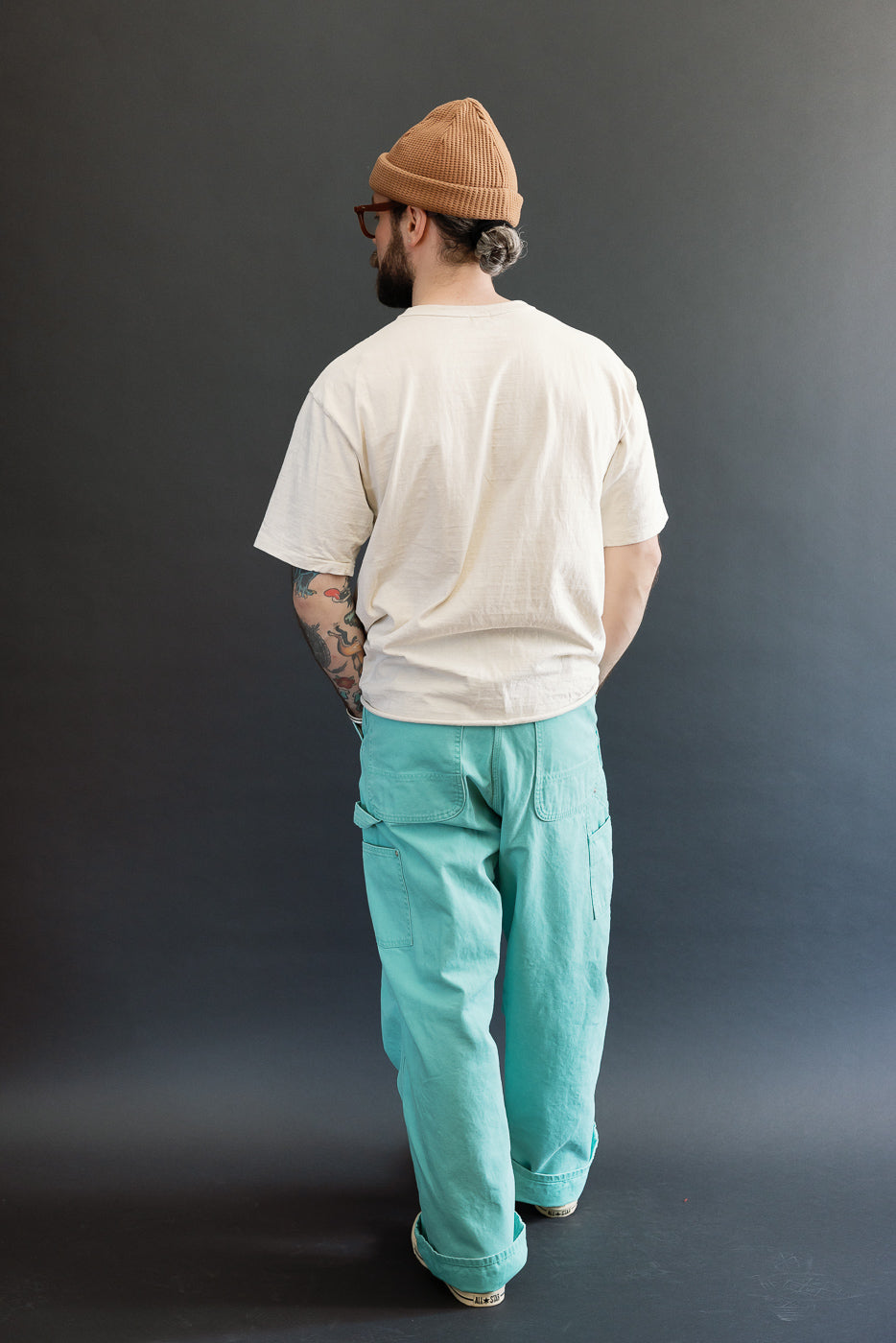Man wearing 01-5328-39SK Dad's Fit Painter's Pants Canvas - Turquoise, with loose fit and brass UFO rivet detail, styled with a cream T-shirt and tan beanie, showcasing workwear elegance.
