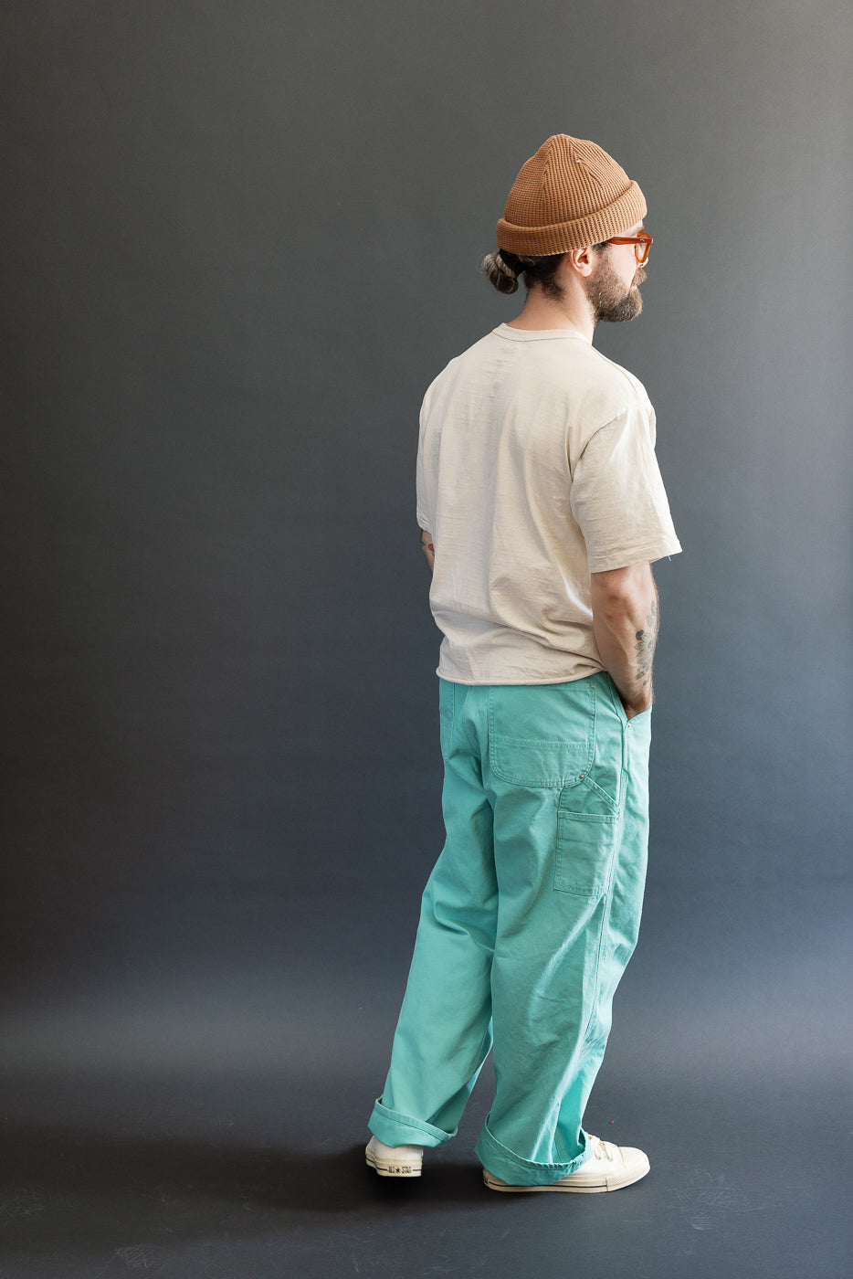 A man models the 01-5328-39SK Dad's Fit Painter's Pants Canvas - Turquoise, showcasing their loose fit, patch pockets, and brass UFO rivet details, paired with a beige T-shirt and camel-brown beanie.
