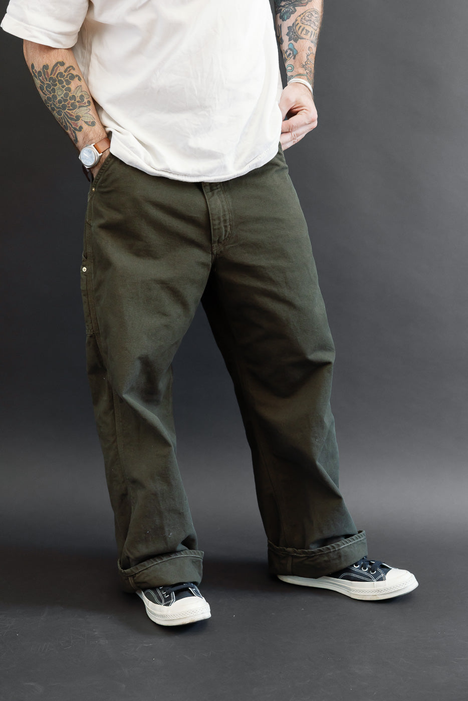 Full body front view of a male model with an athletic build wearing olive green casual trousers and black canvas sneakers.