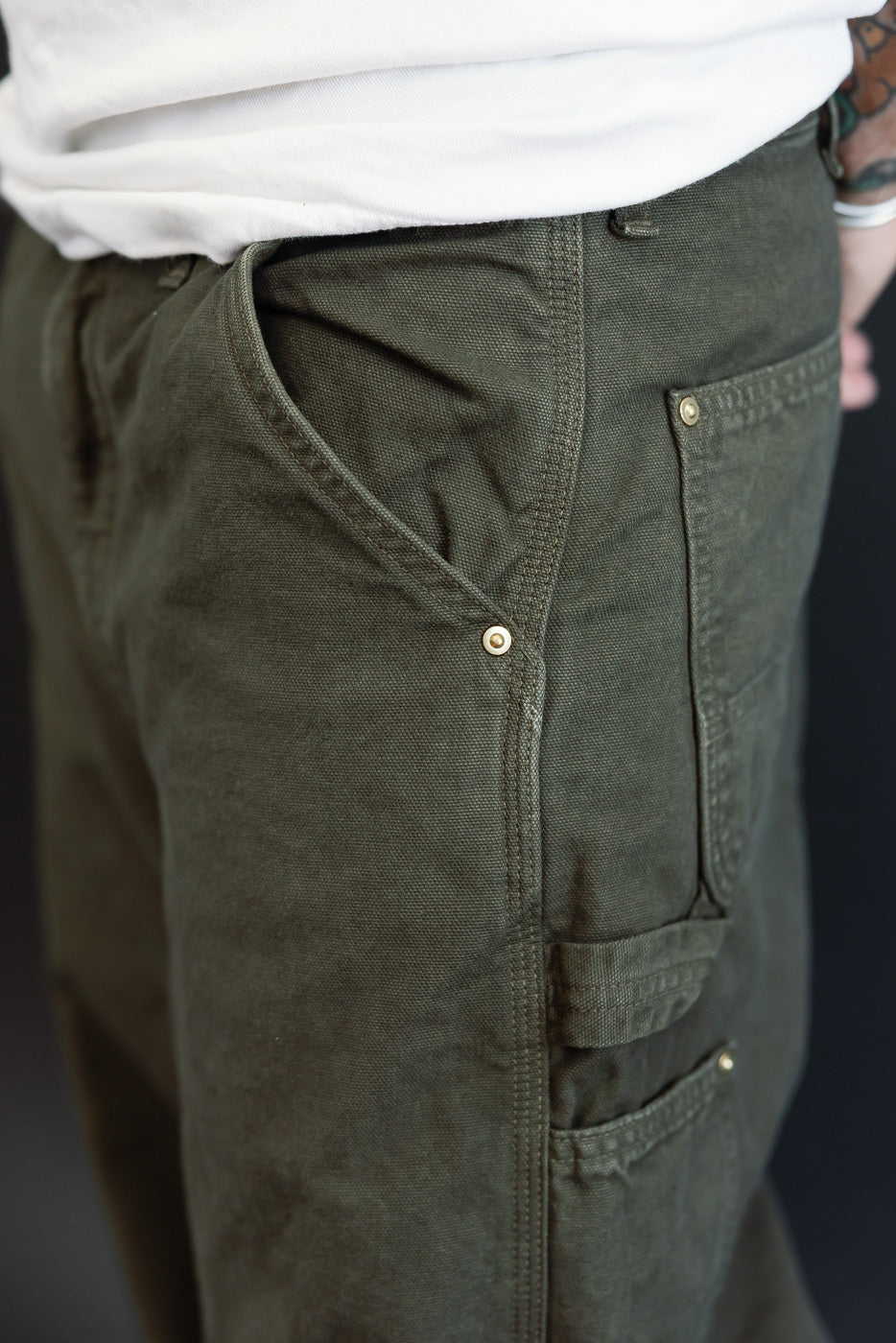 Close-up side view of olive green canvas utility pants showing pocket detail and metal rivets.