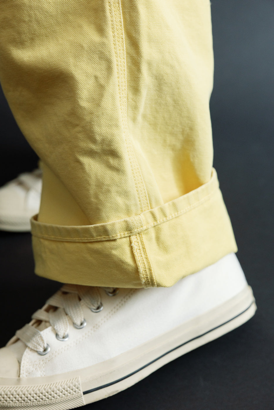 Close-up of male model's lower leg in light yellow cotton pants with cuffed hem, paired with white sneakers