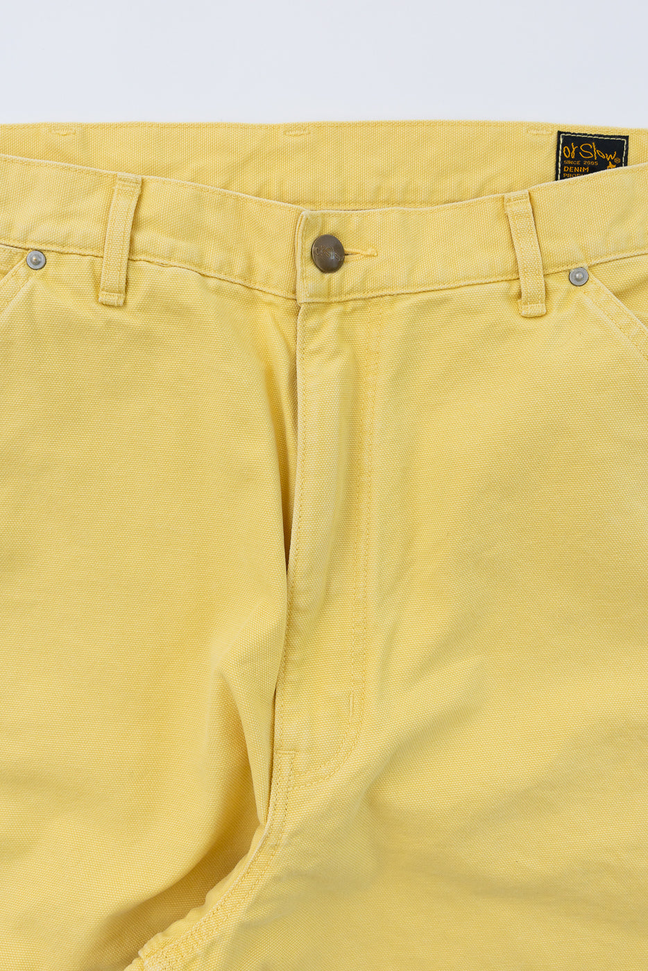 Close-up front view of yellow cotton twill pants with button fly, metal rivets, and brand tag at waistband