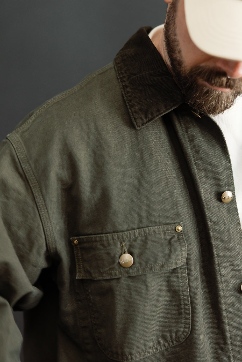 Close-up front view of an olive green canvas jacket featuring a corduroy collar and chest pocket.
