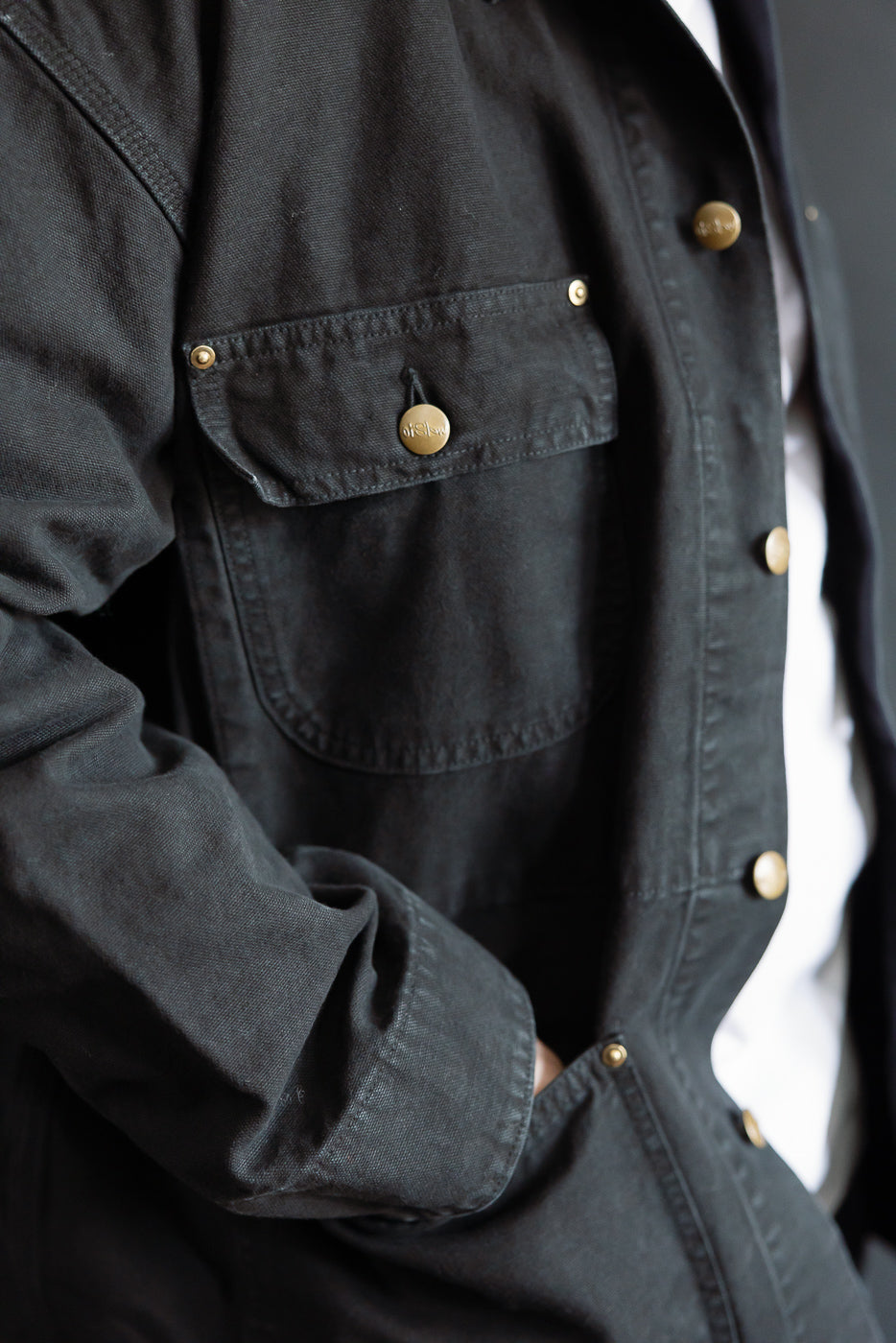 Close-up of male model's left chest and sleeve wearing black Orslow coverall jacket, brass snap buttons, front pocket detail. Model is 5'9