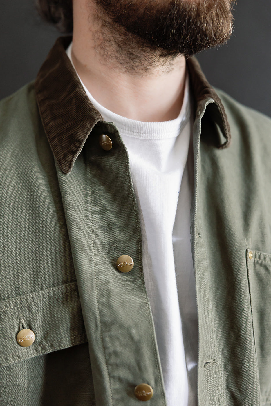 Close-up of front upper orSlow army green coverall jacket on male model, olive fabric, corduroy collar, brass snap buttons, Dant Indianapolis.