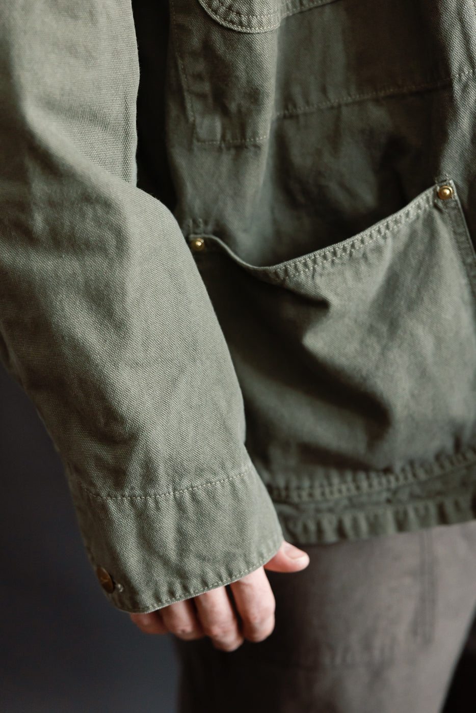 Close-up of male model's arm and front lower pocket on orSlow army green coverall jacket, showing fabric texture and brass rivet details.