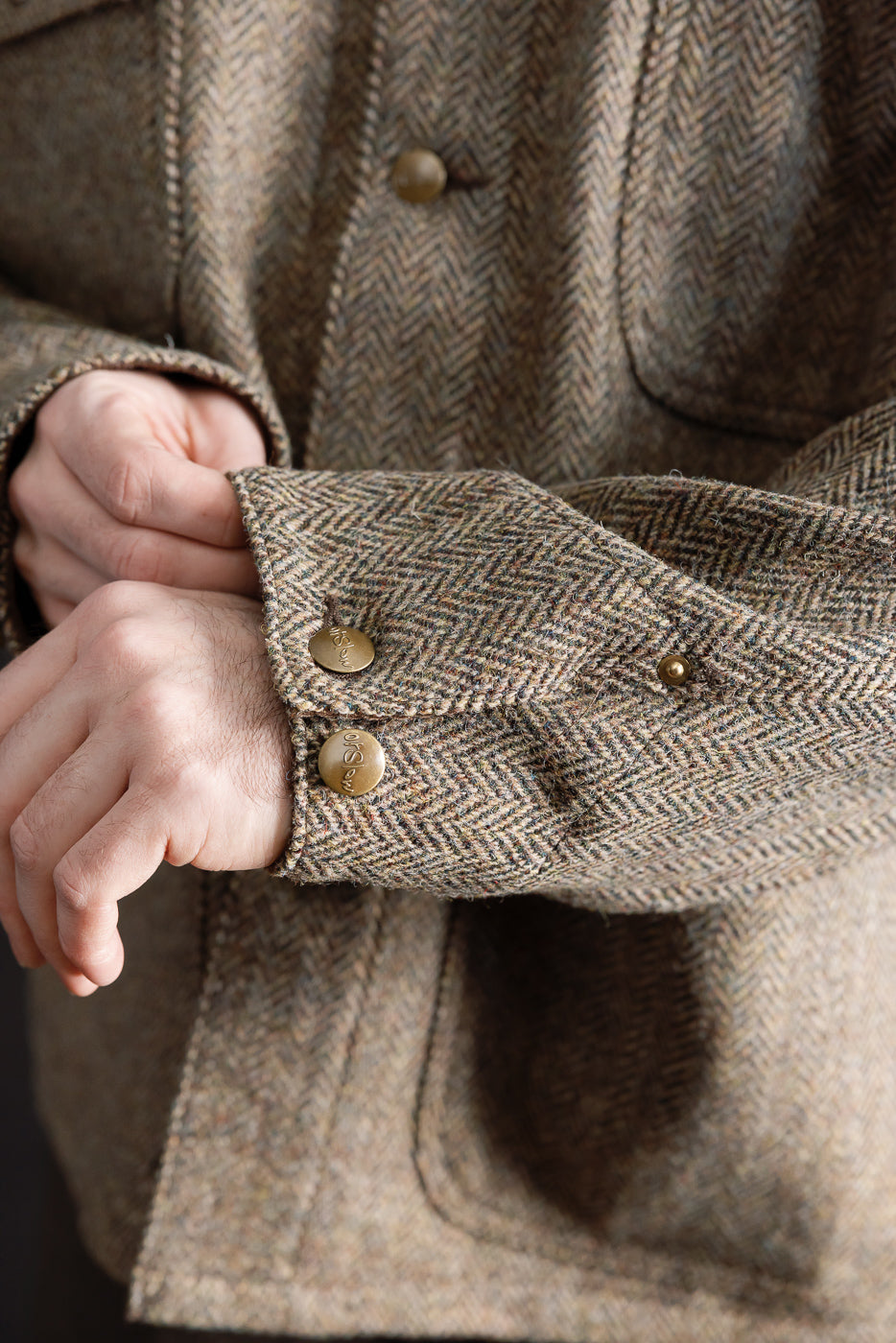 Close-up of male model's wrist on orSlow Harris Tweed coverall jacket, showing brass cuff buttons, sleeve texture, and chest pocket detail in khaki herringbone. Model is 5'9