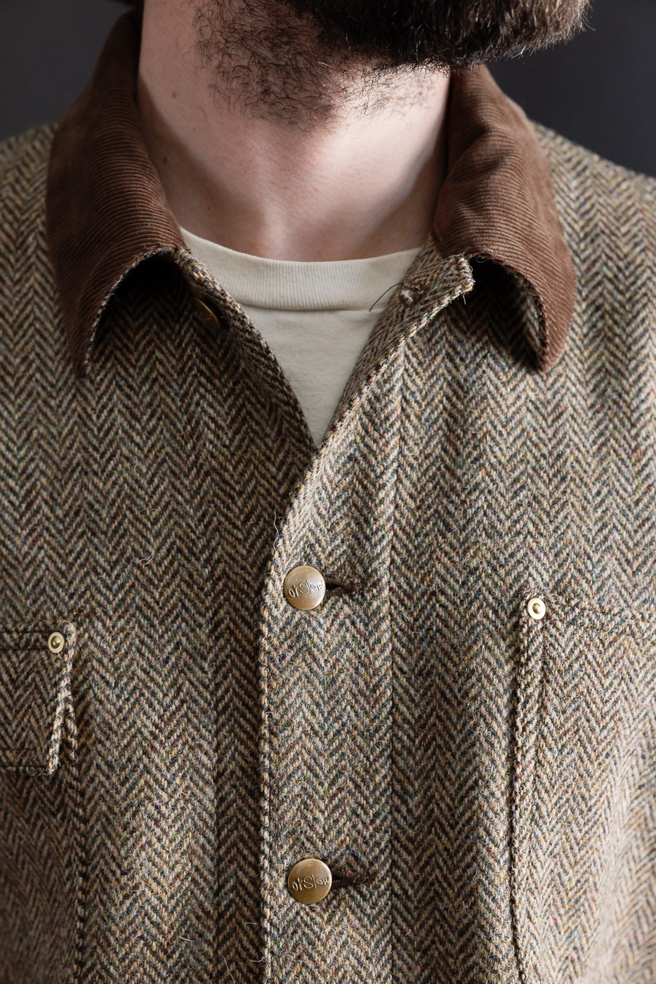 Close-up of front chest area of Orslow Harris Tweed khaki coverall jacket on male model, showing brown corduroy collar, herringbone wool texture, branded buttons, and pockets. Model is 5'9