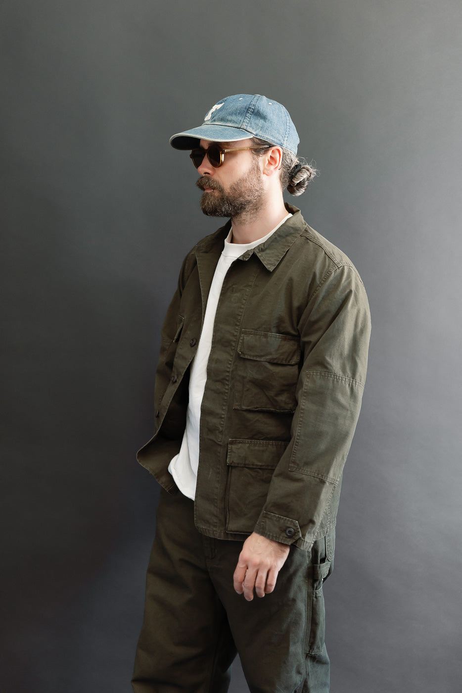 Full body profile view of a male model with an athletic build wearing an olive green utility jacket and matching trousers.