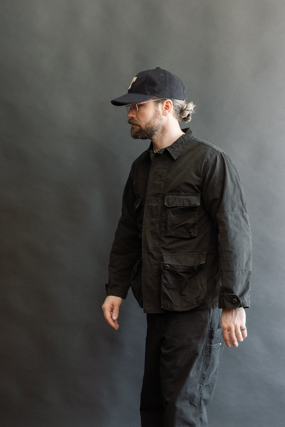 Man in black 01-6110-61S Ripstop BDU Jacket - Black Stone, featuring flap pockets and a utilitarian design, stands in profile. Styled with matching trousers, showcasing premium menswear from Dant.