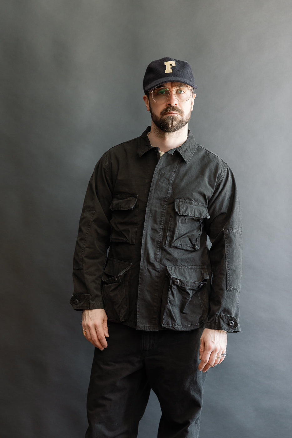 Man wearing the 01-6110-61S Ripstop BDU Jacket in Black Stone, featuring multiple flap pockets and a utilitarian design, styled with matching trousers and a navy cap, embodying a minimalist, military-inspired look.