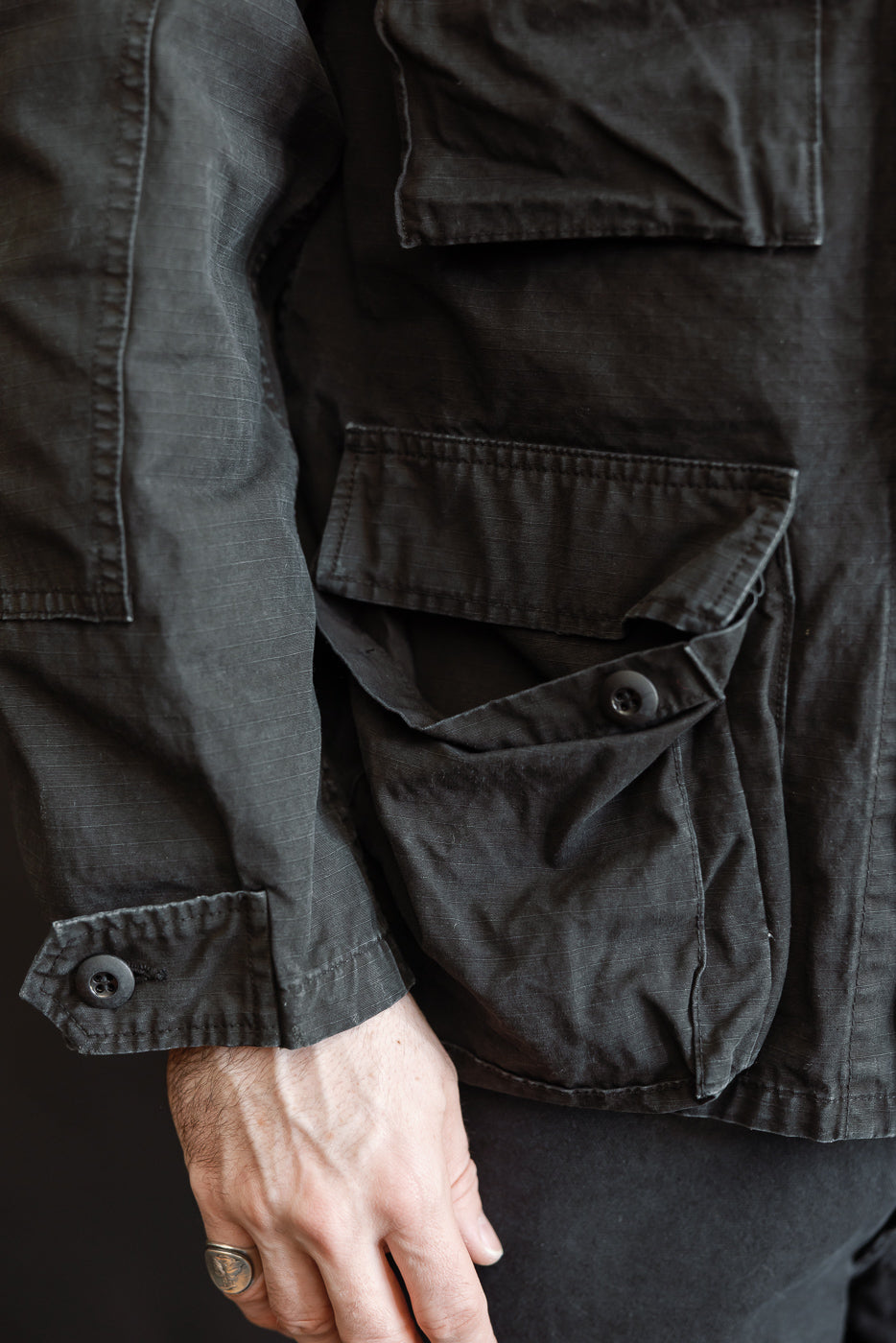 Close-up of the 01-6110-61S - Ripstop BDU Jacket - Black Stone, showcasing a flap pocket, buttoned cuff, and a hand wearing a silver ring, highlighting the jacket's utility design and ripstop texture.