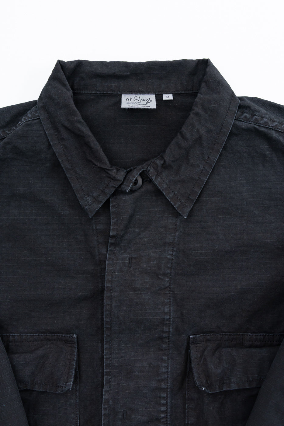 Close-up of 01-6110-61S - Ripstop BDU Jacket - Black Stone, featuring a black button-up design, pointed collar, hidden-placket, flap chest pockets, and orSlow – Made in Japan label, highlighting its military-inspired style.
