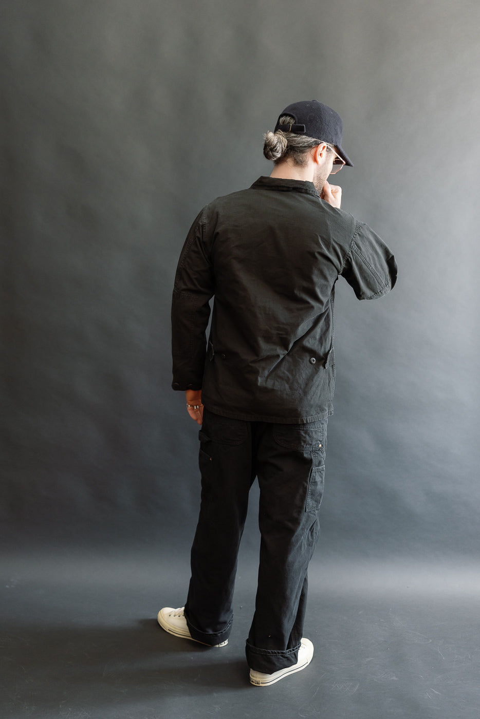 A person stands wearing the 01-6110-61S Ripstop BDU Jacket - Black Stone, showcasing its military-inspired design with four front patch pockets and adjustable cuffs, paired with black cargo pants and white sneakers.