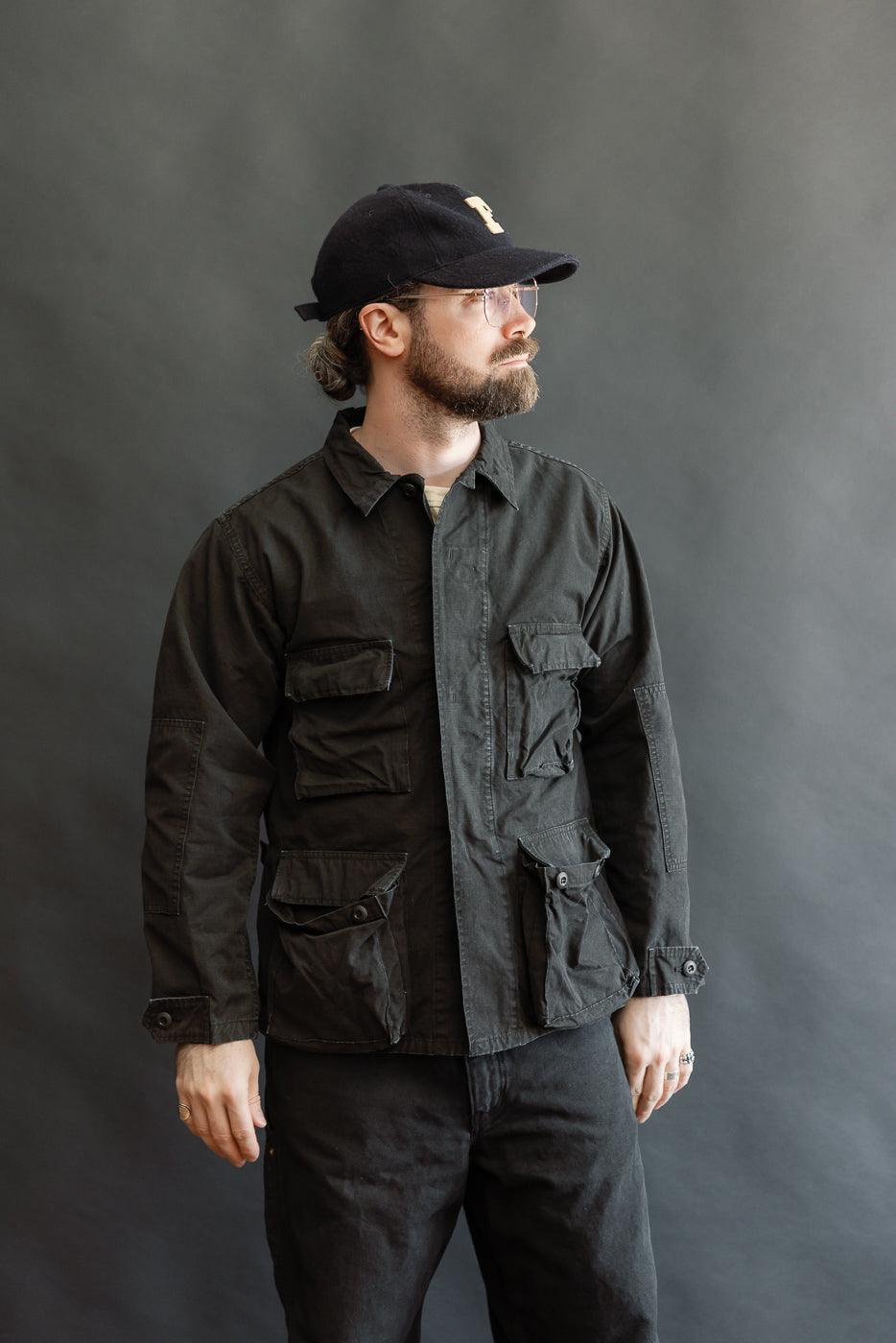 A bearded man models the 01-6110-61S - Ripstop BDU Jacket - Black Stone, showcasing its military-inspired design with multiple flap pockets, clear-framed glasses, and a black cap, embodying Dant's premium menswear style.