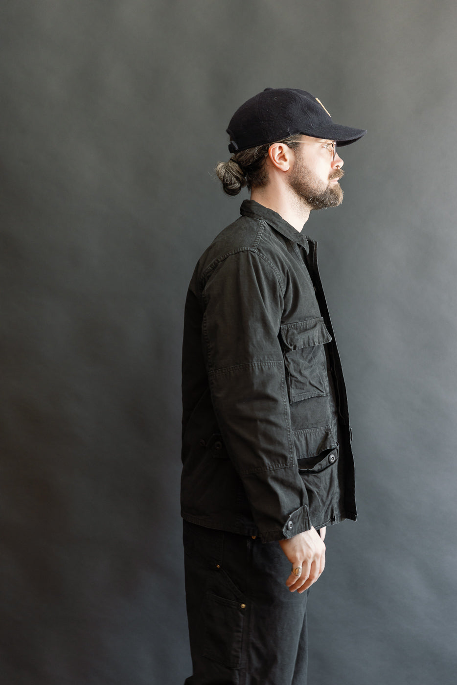 Bearded man in a black stonewashed 01-6110-61S Ripstop BDU Jacket, featuring a military design with flap pockets and replica buttons, posed in profile against a gray backdrop.