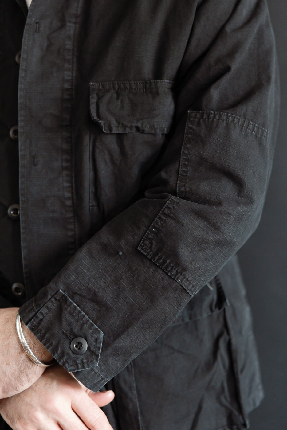Close-up of black ripstop BDU jacket sleeve and front pocket on male model, showing buttoned cuff, flap pocket, and fabric texture. Model is 5'9