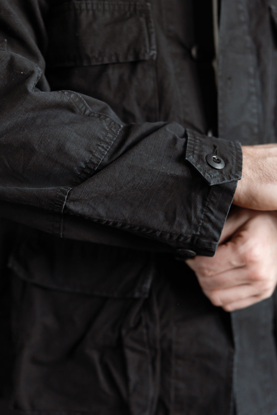Close-up of male model wearing black ripstop BDU jacket, showing sleeve cuff, button, and chest pocket detail, front side. Model is 5'9
